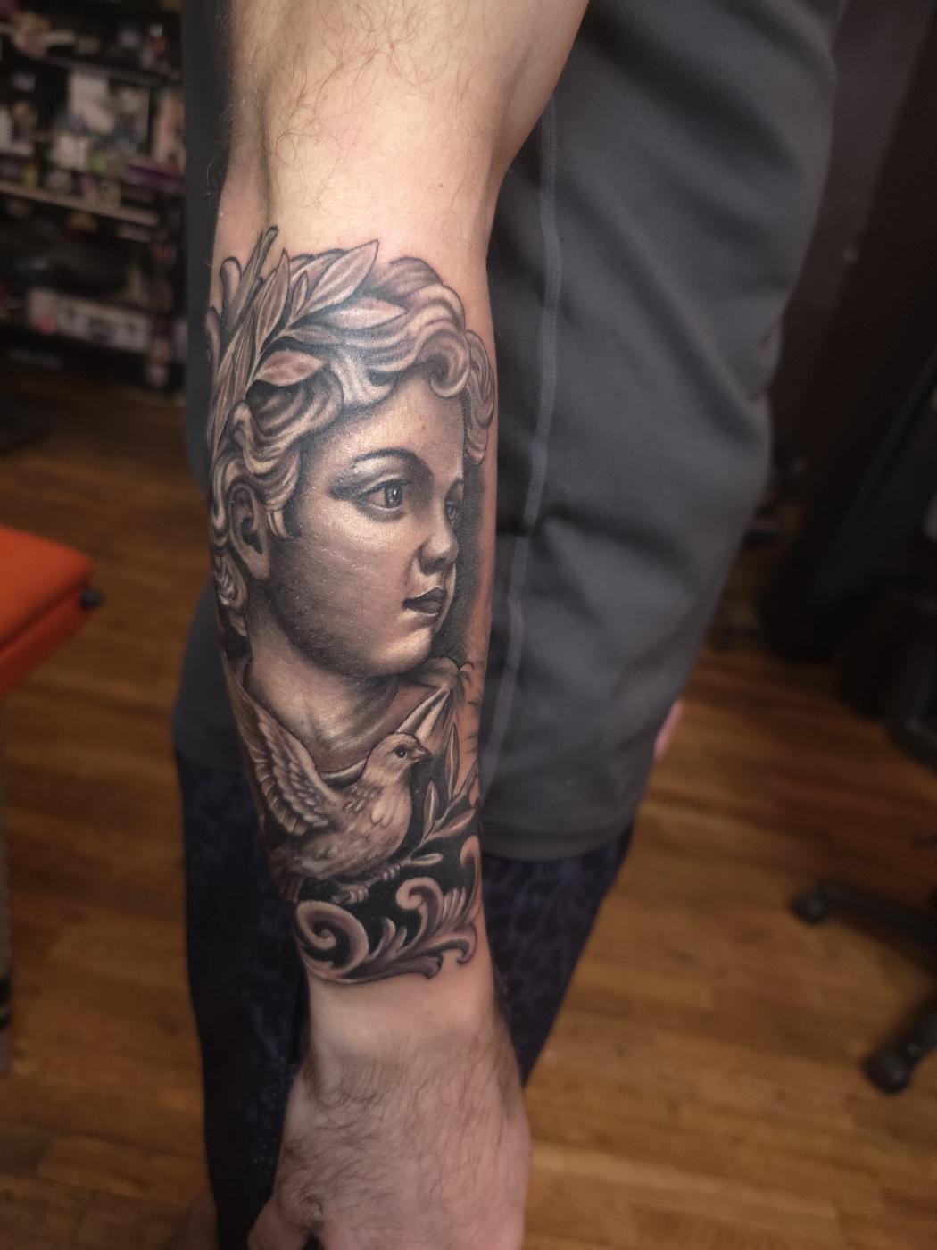 Black and gray portrait with a bird and scrollwork, tattooed on a forearm